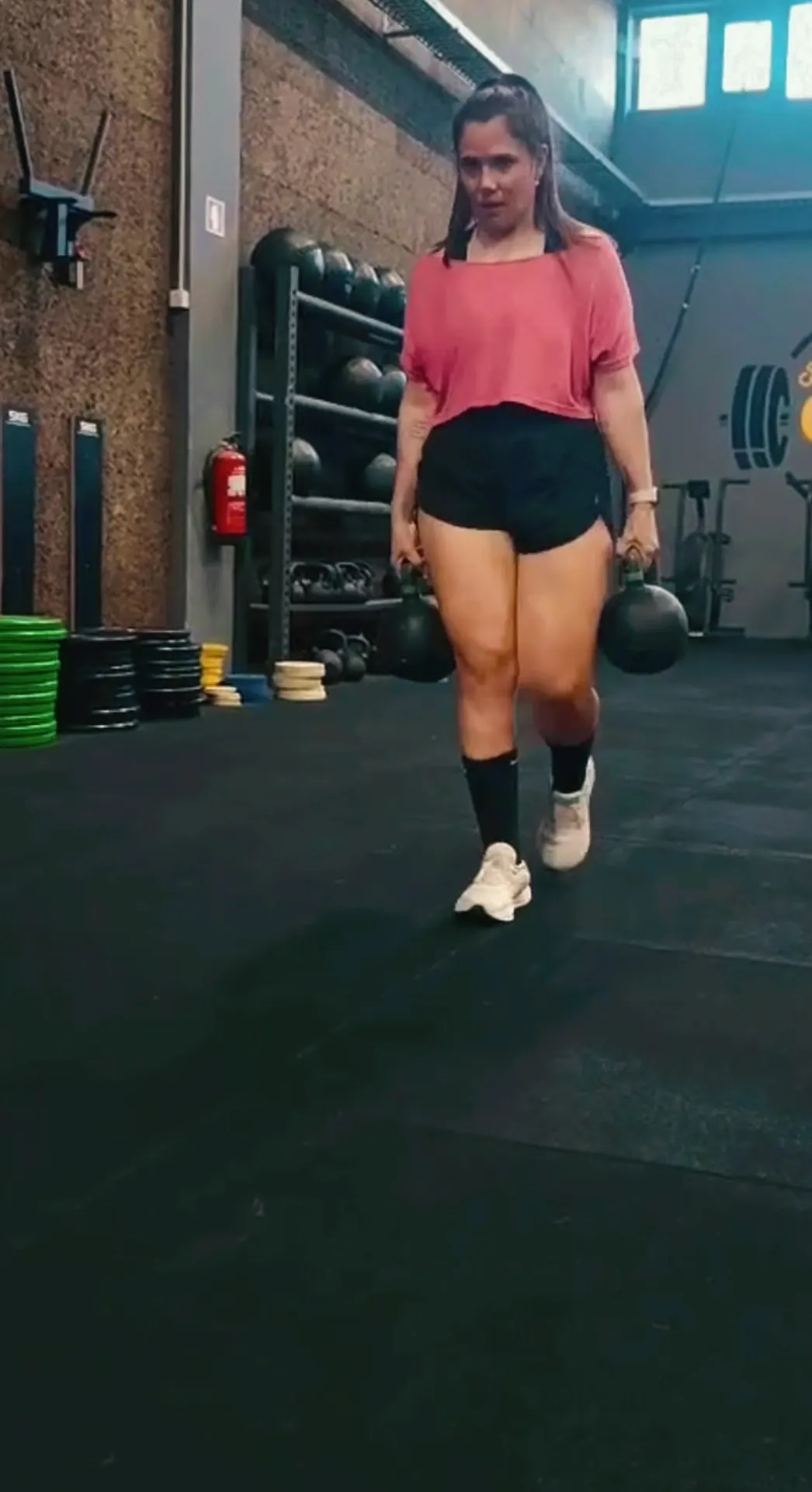 Atleta a caminhar com duas kettlebells no chão de borracha da box